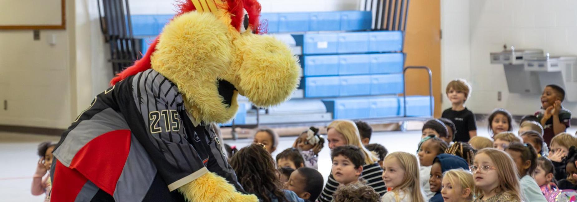 A mascot interacts with a large group of children in a school setting.