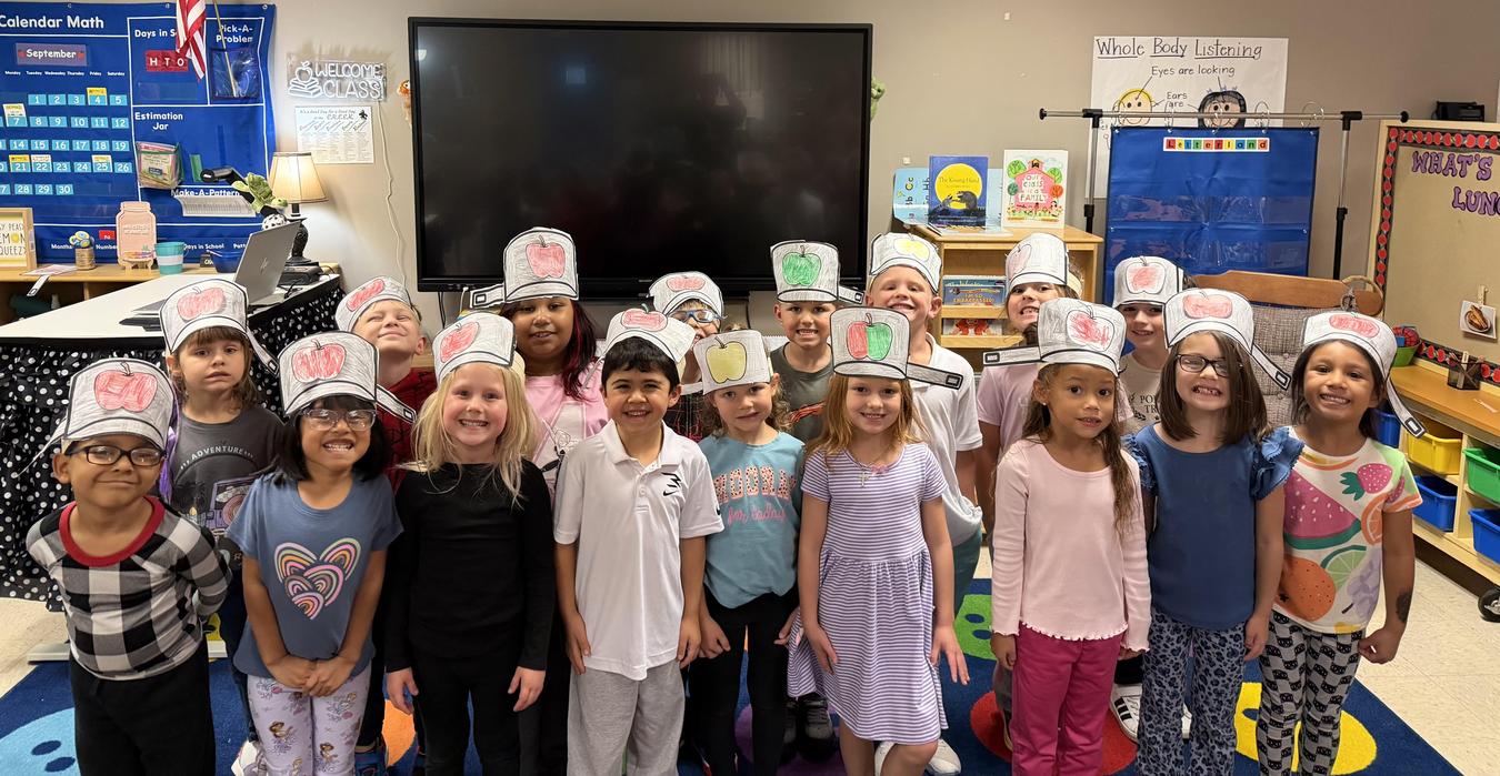 Children in a classroom wearing craft hats with drawings, smiling together.