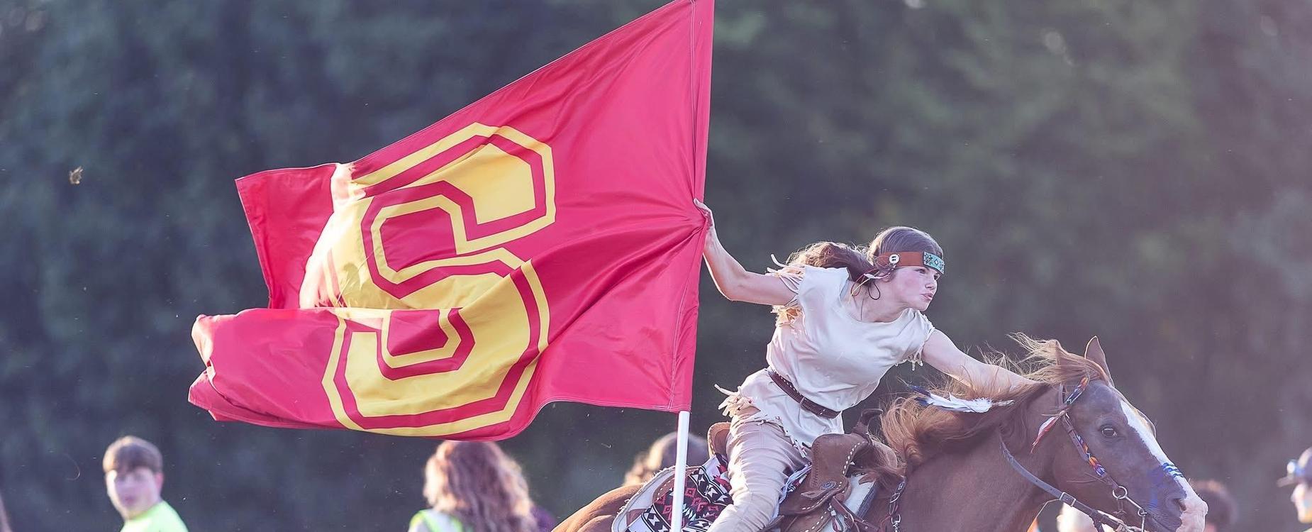 A rider in a cowboy outfit gallops on a horse while waving a large red flag.