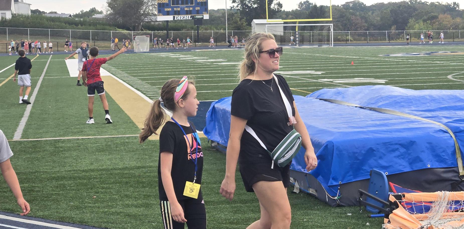 A girl and a woman walking together on a field with spectators in the background.
