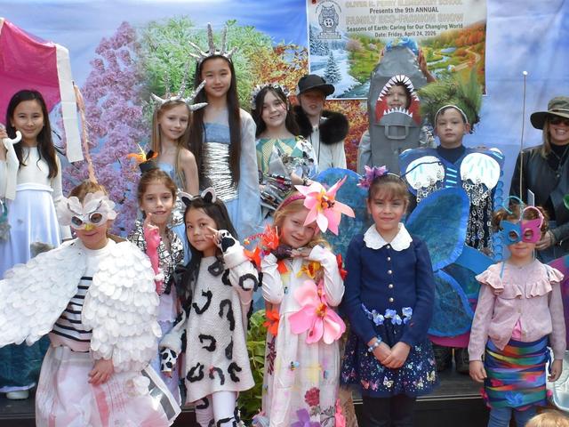 A diverse group of children in costumes stands on stage, posing with creative outfits.