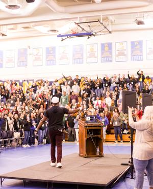A crowd of people cheer during an event, with a speaker on stage.
