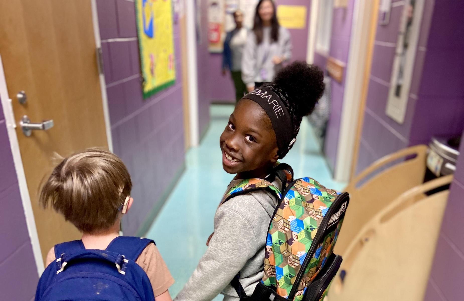 Smiling girl with backpack walking down a school hallway with a boy.