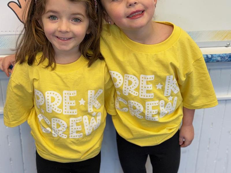 Two girls in yellow shirts with 'PRE-K CREW' text smiling together.