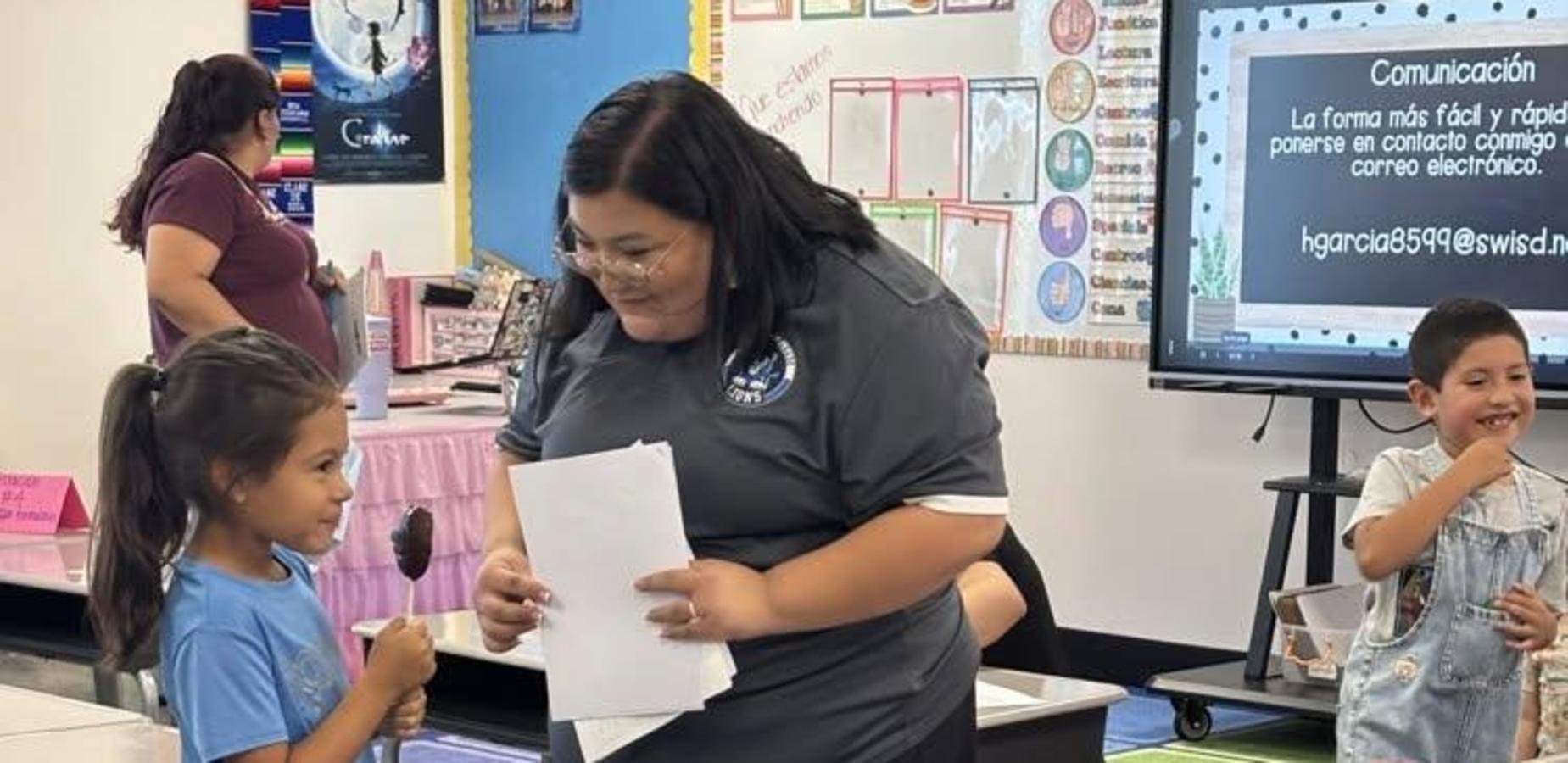 Adult interacts with a child in a classroom, while another child smiles nearby.
