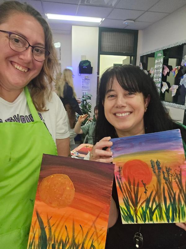 Two women proudly displaying their sunset paintings in an art classroom.