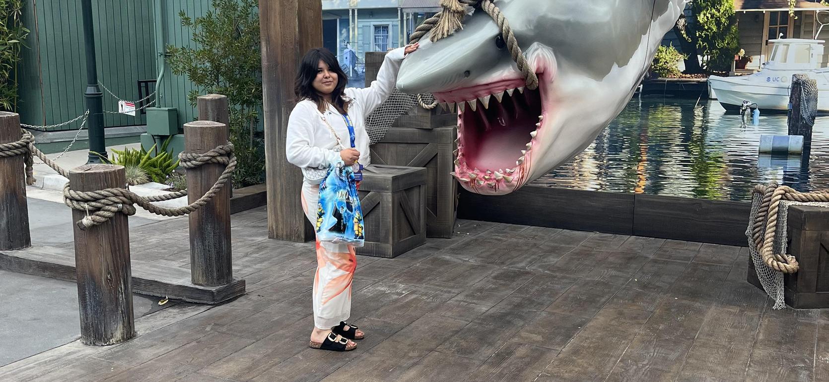 Person posing with a large shark sculpture by a waterfront.