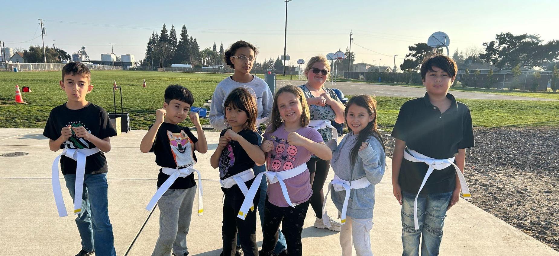 A group of eight children posing with martial arts belts at an outdoor area.