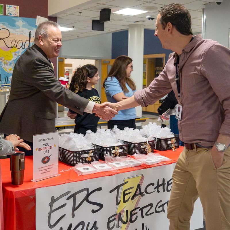 Two men shaking hands at a school event with a table of snacks behind them.