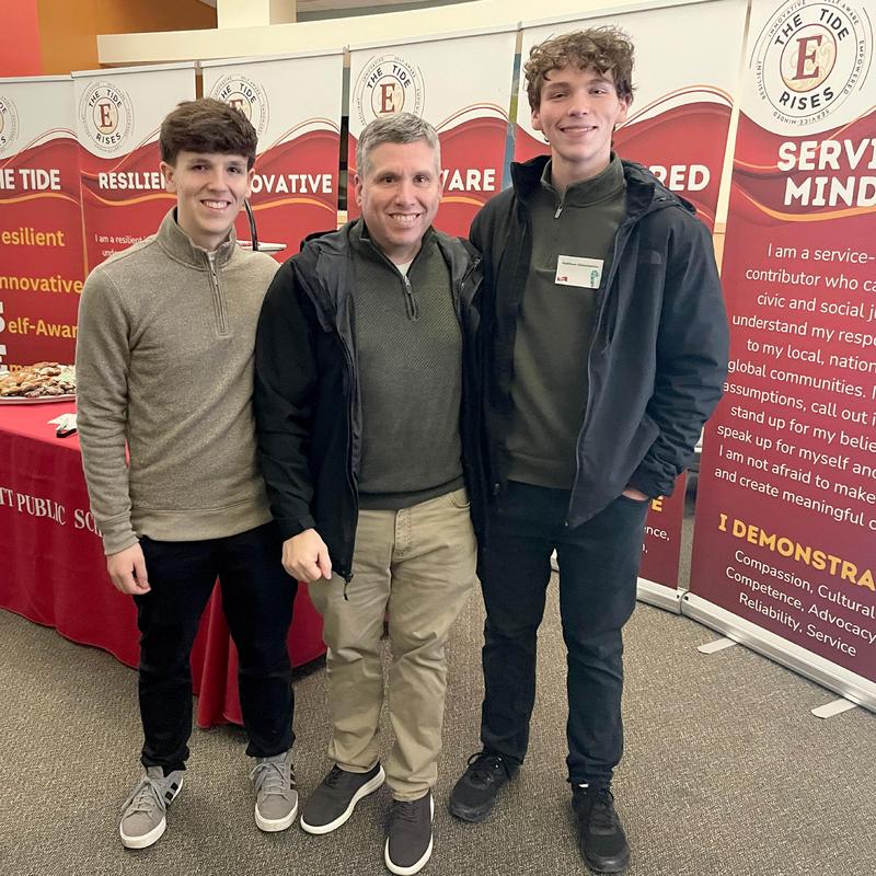 Three men standing together, smiling in a conference setting with banners in the background.