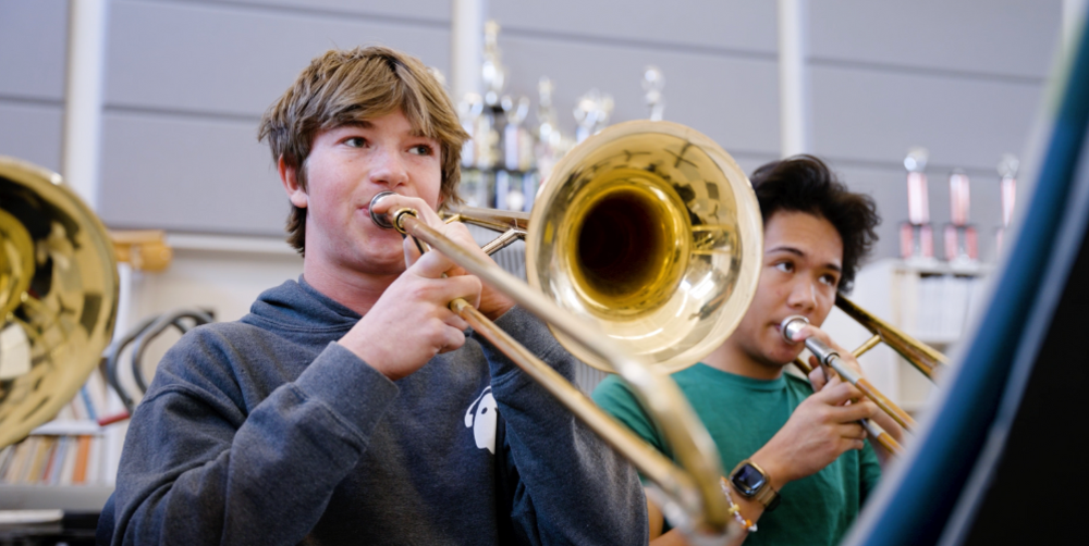 Two students playing trombones in a music classroom, one wearing a hoodie.