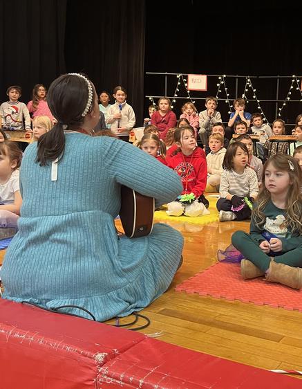 Children sitting on a stage with a performer playing guitar in front of them.