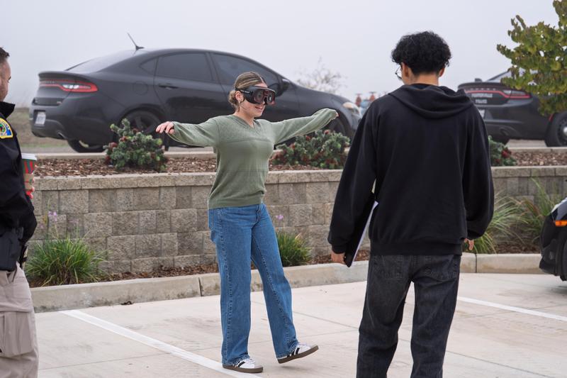 A student wearing impairment goggles attempts a balance test while being observed by a fellow student and a law enforcement officer.