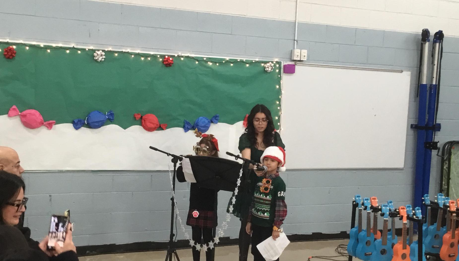 Two children singing at a microphone with an audience capturing the moment on their phones.