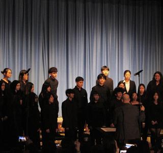 A choir of students performing on stage in dark attire with a conductor.