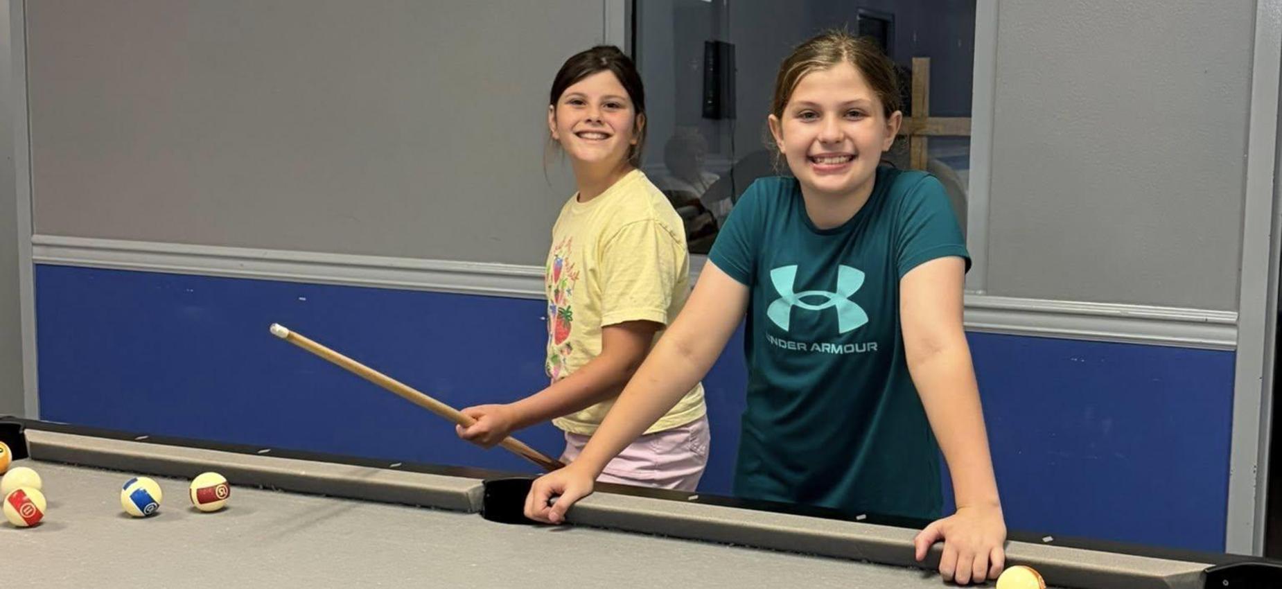 Two girls smiling by a pool table, one holding a cue stick.
