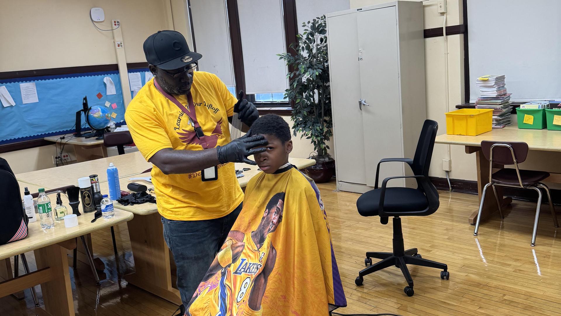 A barber gives a haircut to a young boy wearing a Lakers cape in a classroom setting.