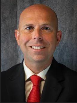 Smiling man in a suit with a red tie against a gray backdrop.