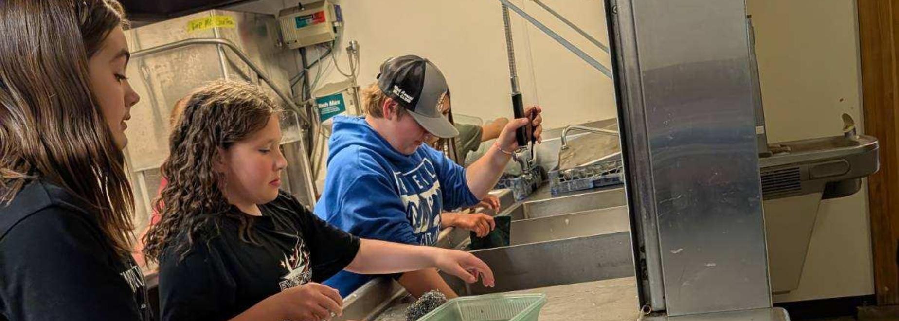 Kids cleaning trays at a kitchen sink with soapy water and focused expressions.