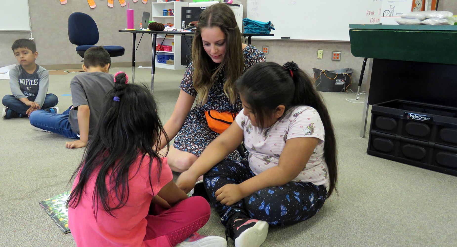 Teacher sits on the floor with two female students as she helps them with a book