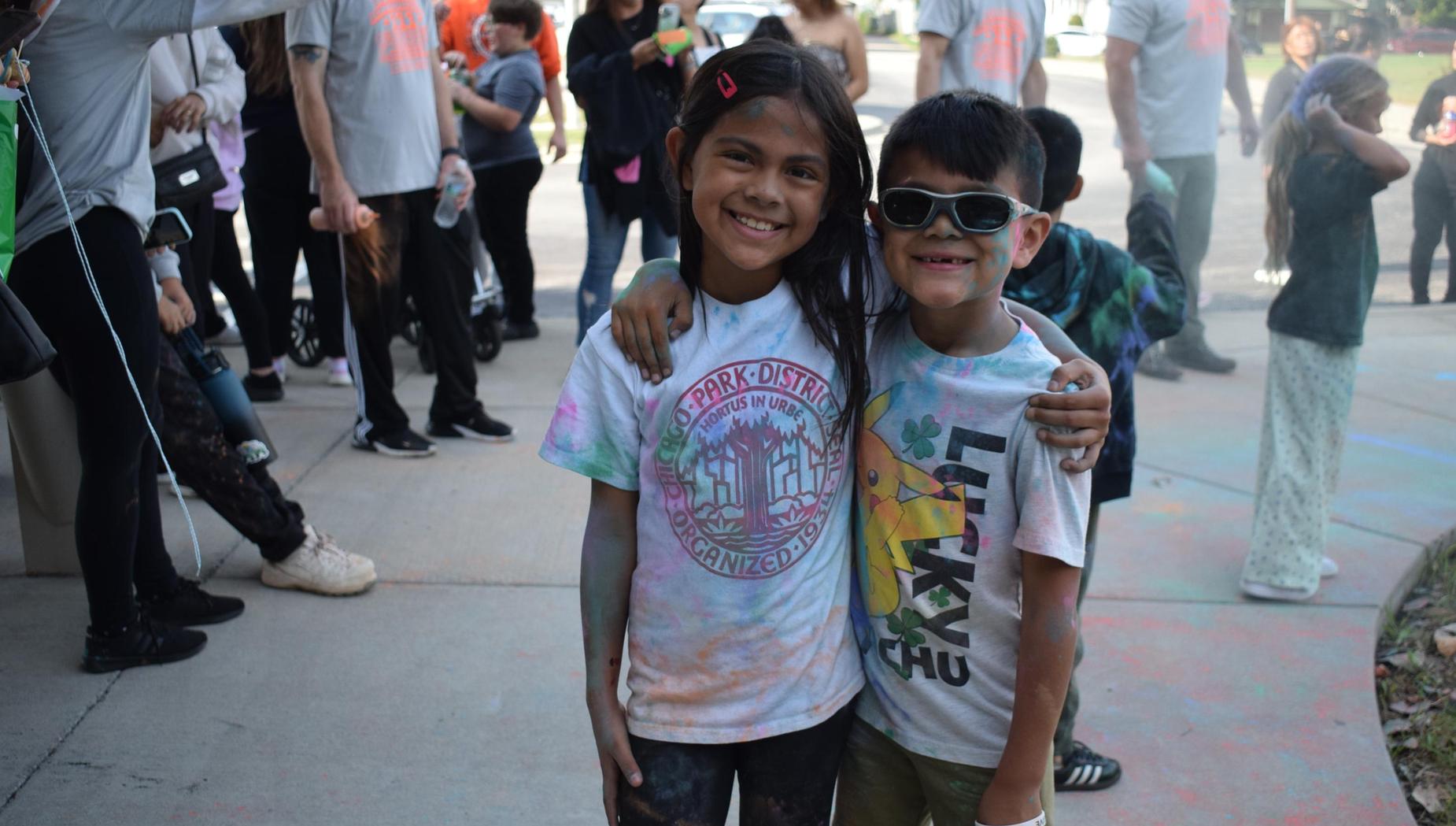 Two smiling children posing together with colorful powder on their clothes at an outdoor gathering.