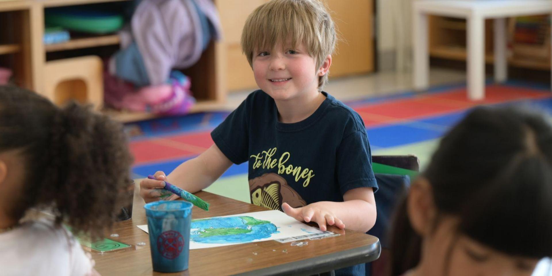 Smiling boy painting a landscape at a table in a colorful classroom.