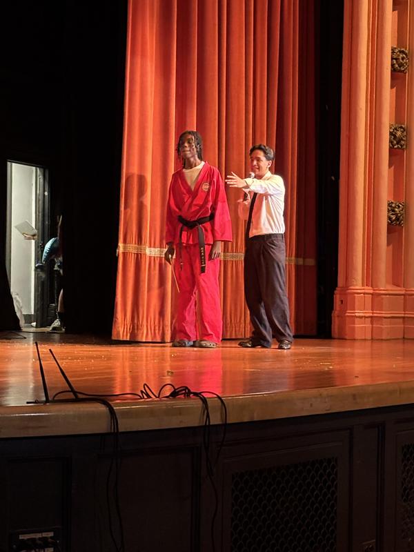Two men on stage, one in a red outfit and another in a suit gesturing toward him.