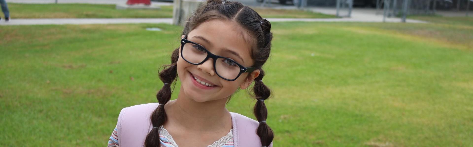 Smiling girl with glasses and braids, standing on green grass.