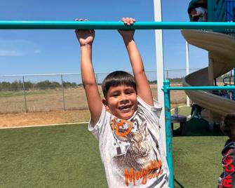 Child hanging on a playground bar, smiling under a blue sky.