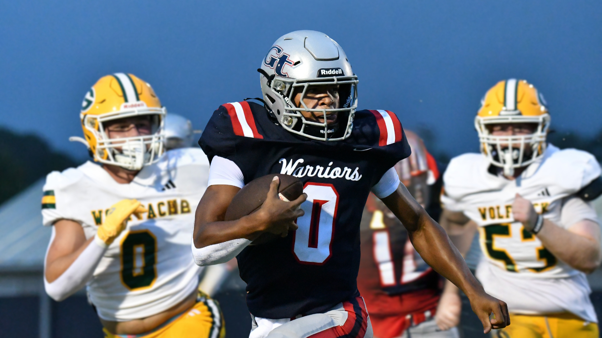 A football player wearing a black jersey with 'Warriors' runs past two defenders.