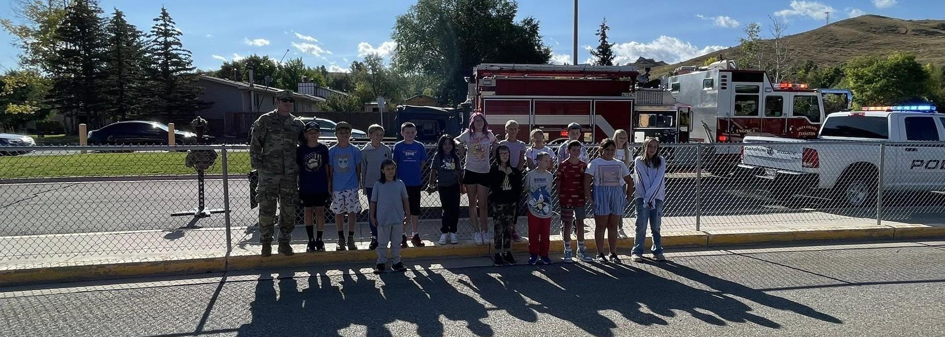A group of children and adults posing in front of emergency vehicles on a sunny day.