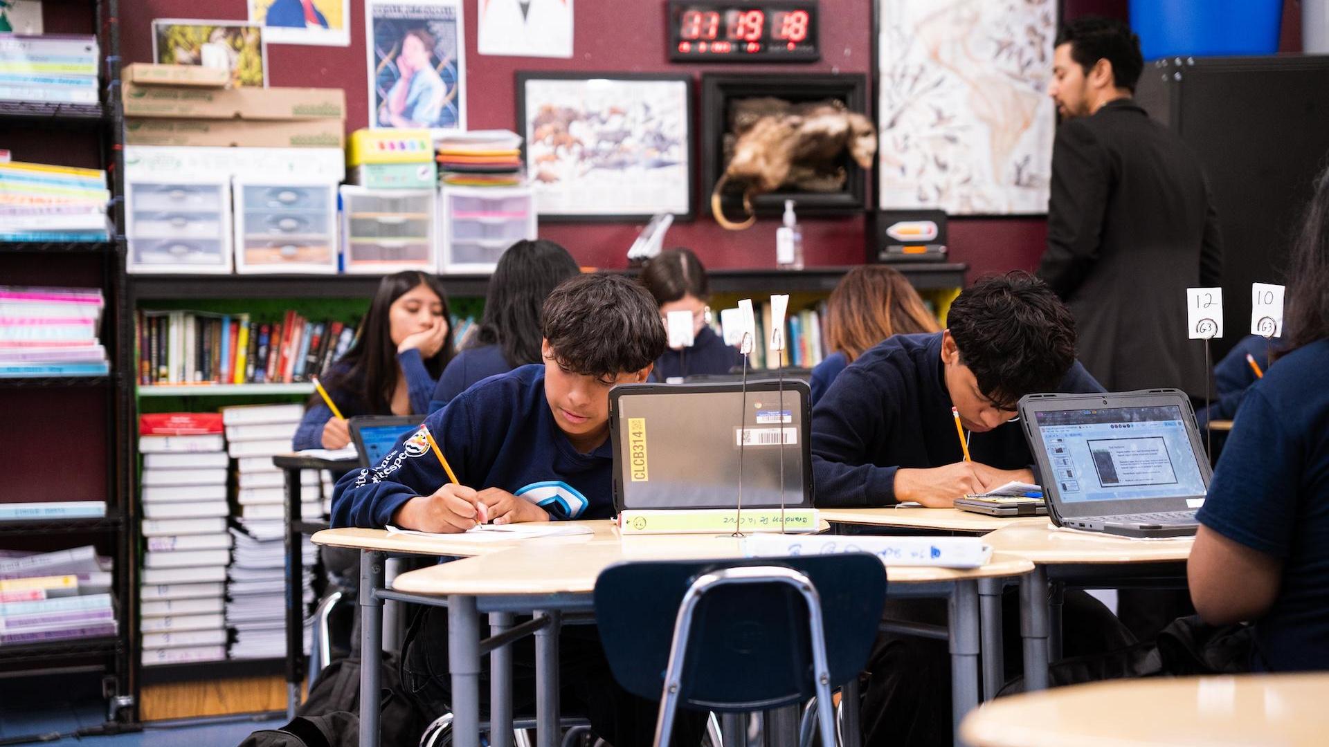 Students seated at desks in a classroom, focused on their laptops and writing.