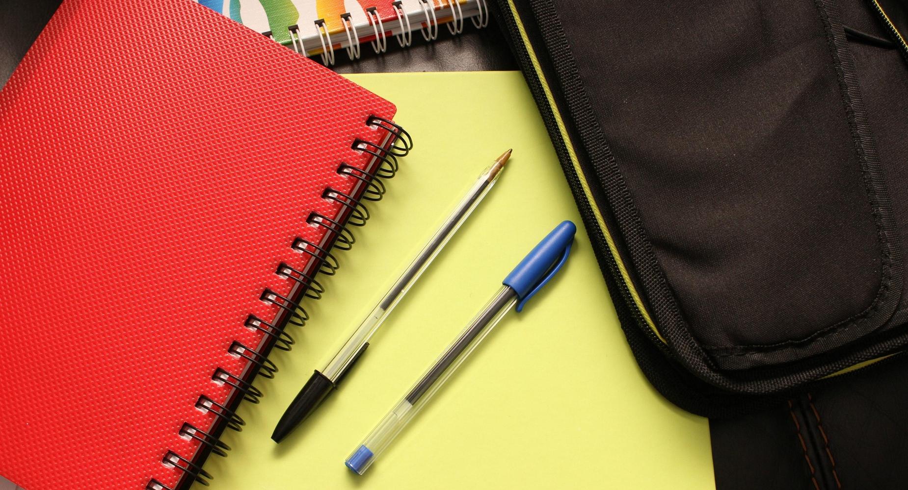 A red notebook, two pens, and a black pencil case on a desk with yellow paper.