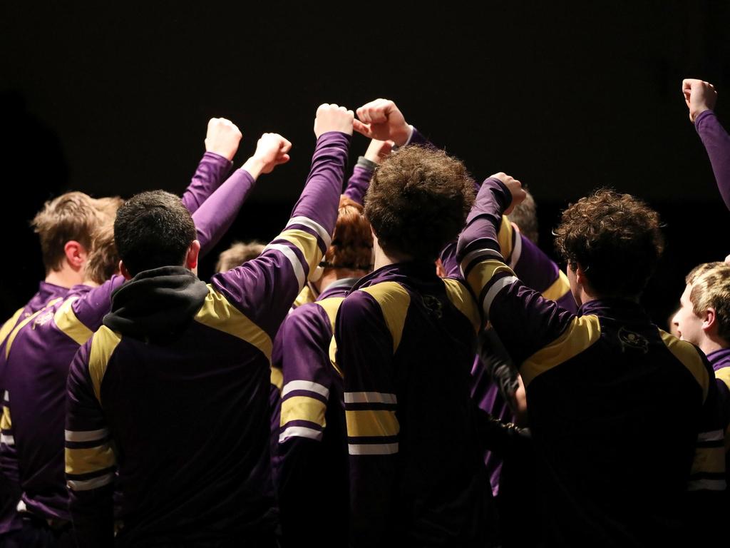 A group athletes wearing purple and gold jerseys standing in a spotlight with their fists raised before a wrestling match.