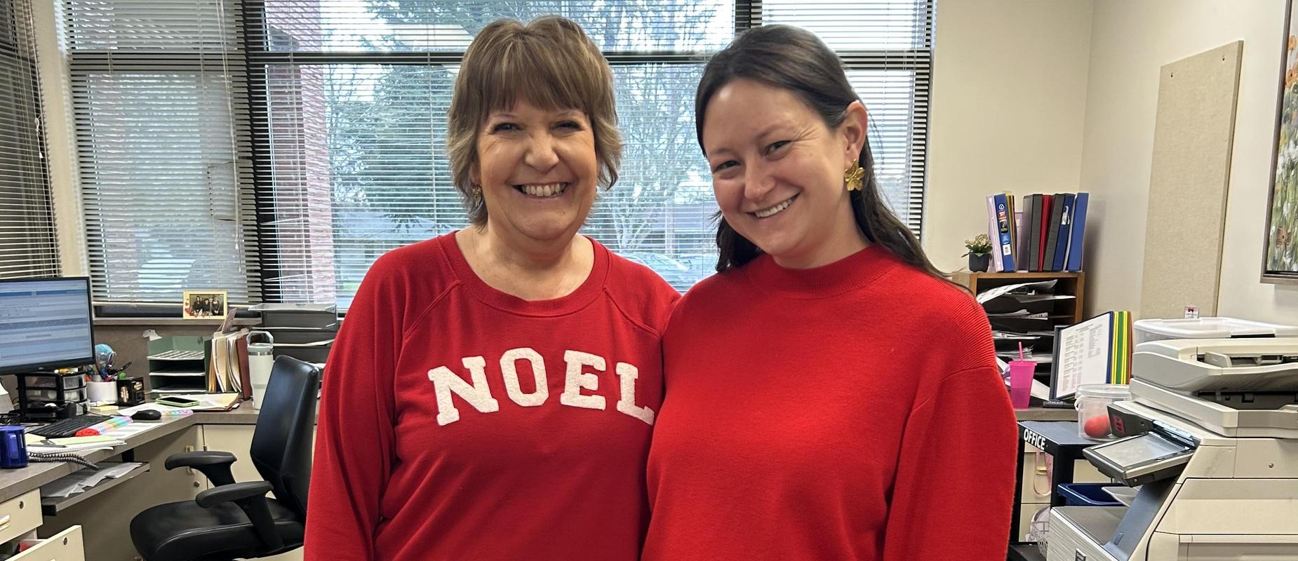 Two women wearing red sweaters standing together in an office setting.