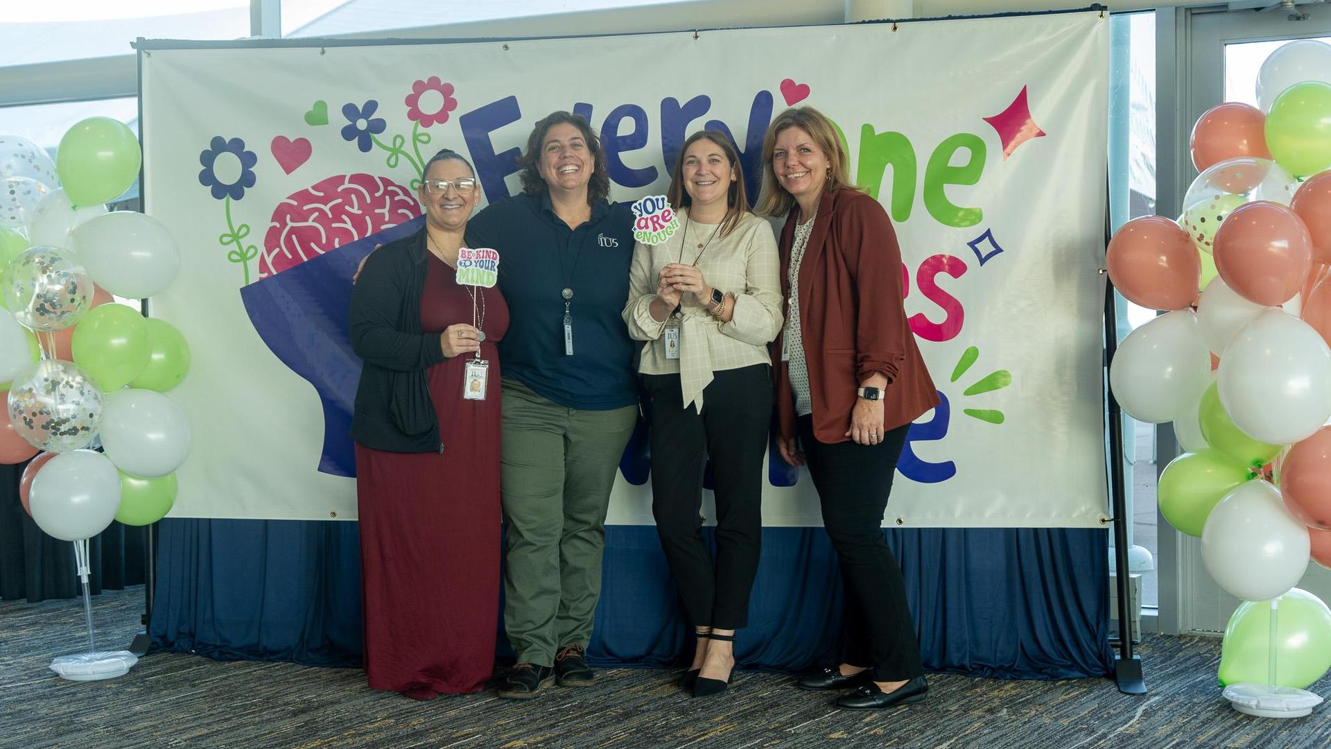 Four women, IU5 staff, pose with props with positive phrases in front of a colorful banner and balloons at a student mental health symposium.