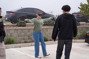 A student wearing impairment goggles attempts a balance test while being observed by a fellow student and a law enforcement officer.