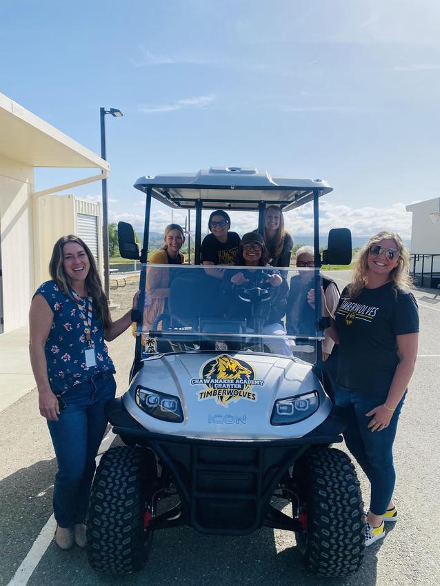 Group of smiling women and one man posing with a golf cart in daylight.