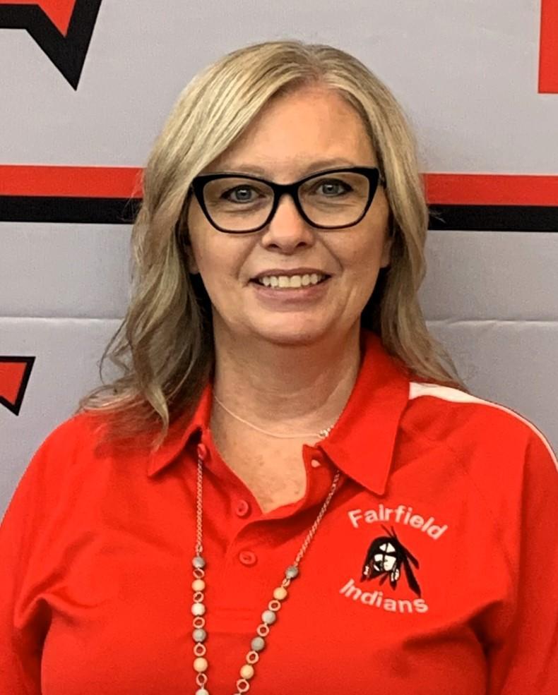 Woman with glasses wearing a red shirt with a school logo and smiling.