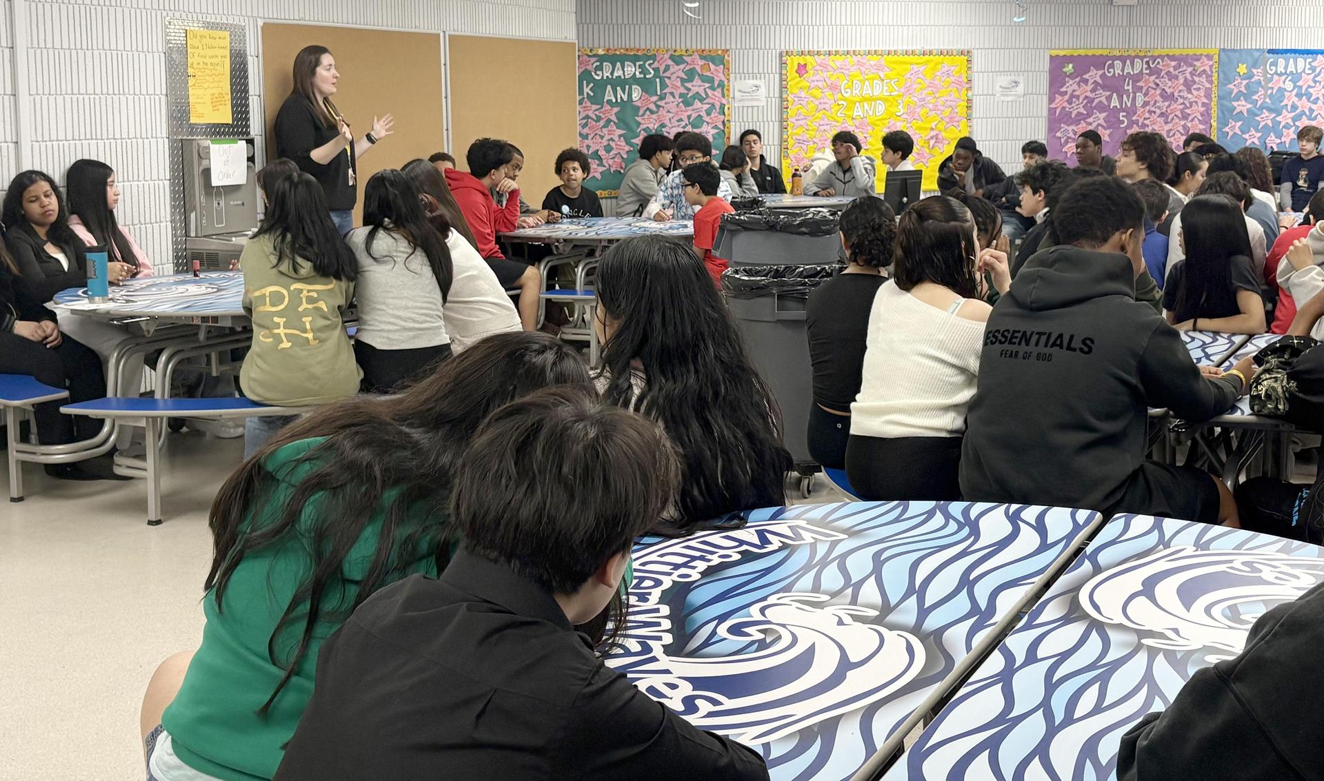 Students gathered in a cafeteria with a speaker presenting at the front.