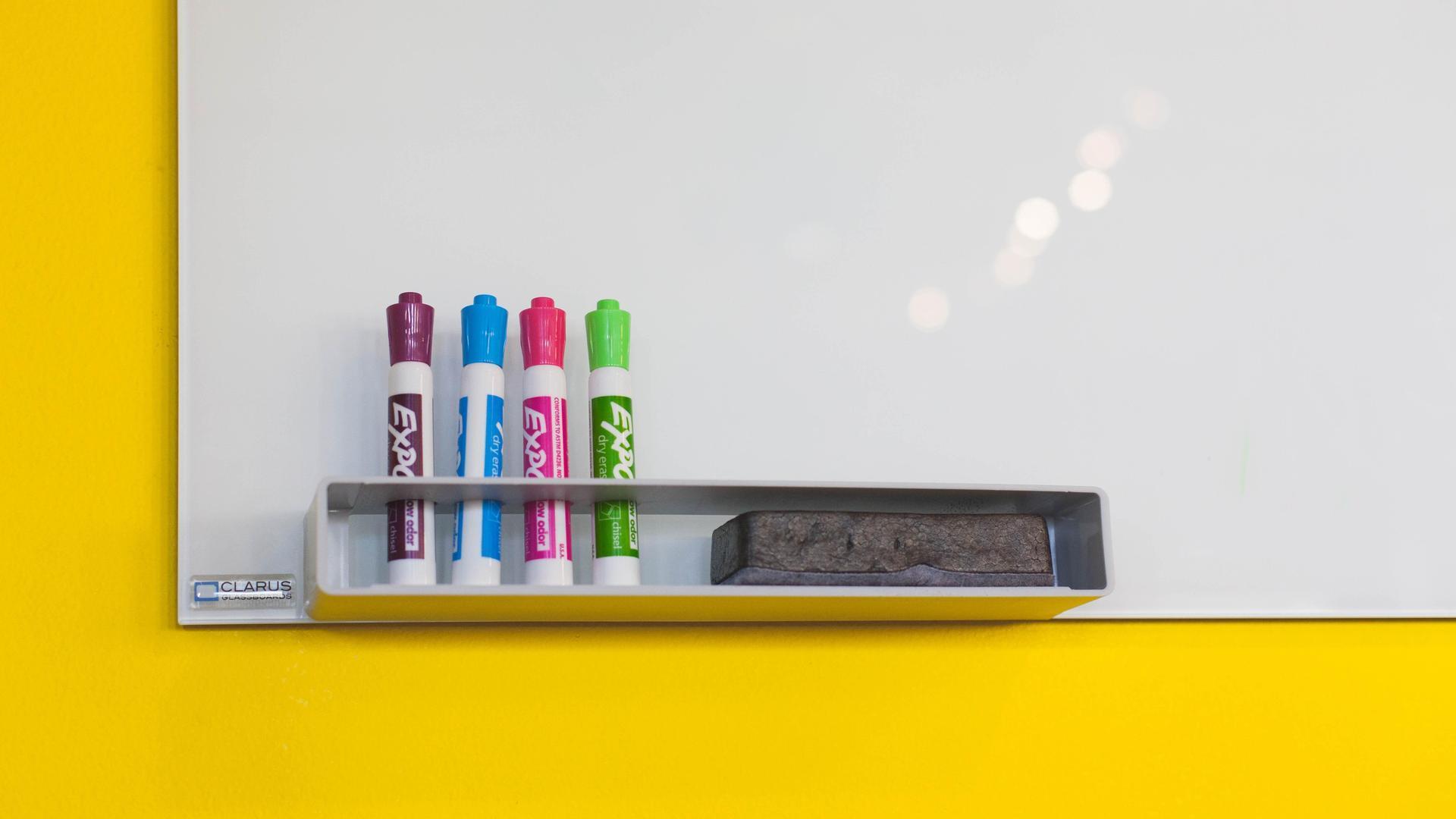 A whiteboard with colorful markers and an eraser on a yellow wall.