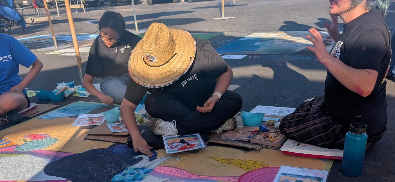 Artists color a large chalk mural on the ground, surrounded by tools and sketches.
