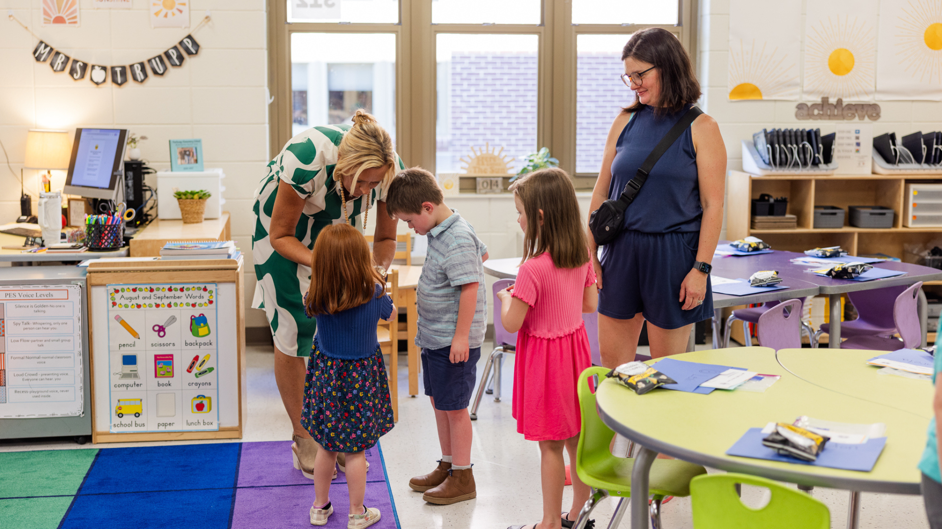 Students greet their teacher in a colorful classroom filled with learning materials.