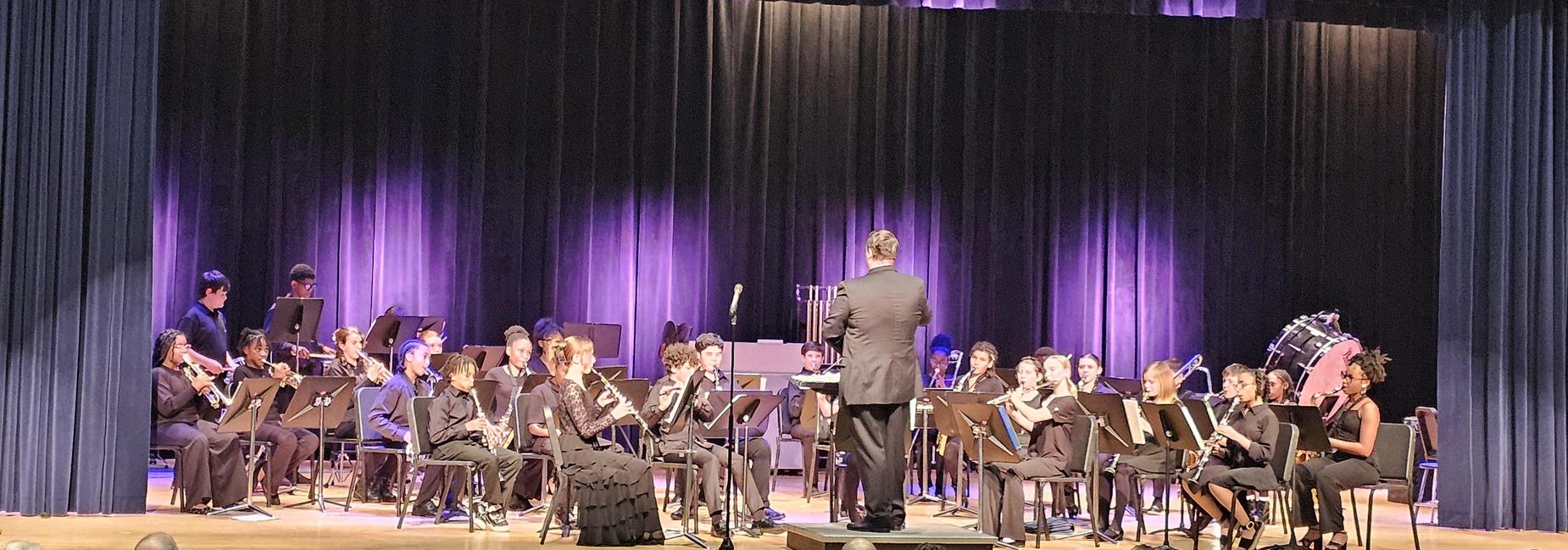 A band performing on stage with a conductor and various instruments under purple lighting.