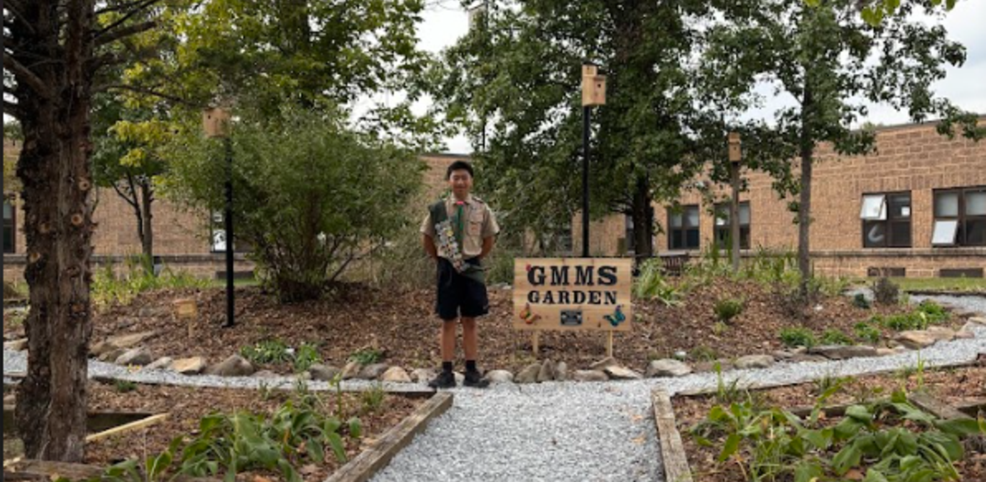A boy stands by the GMMS Garden sign surrounded by plants and trees.