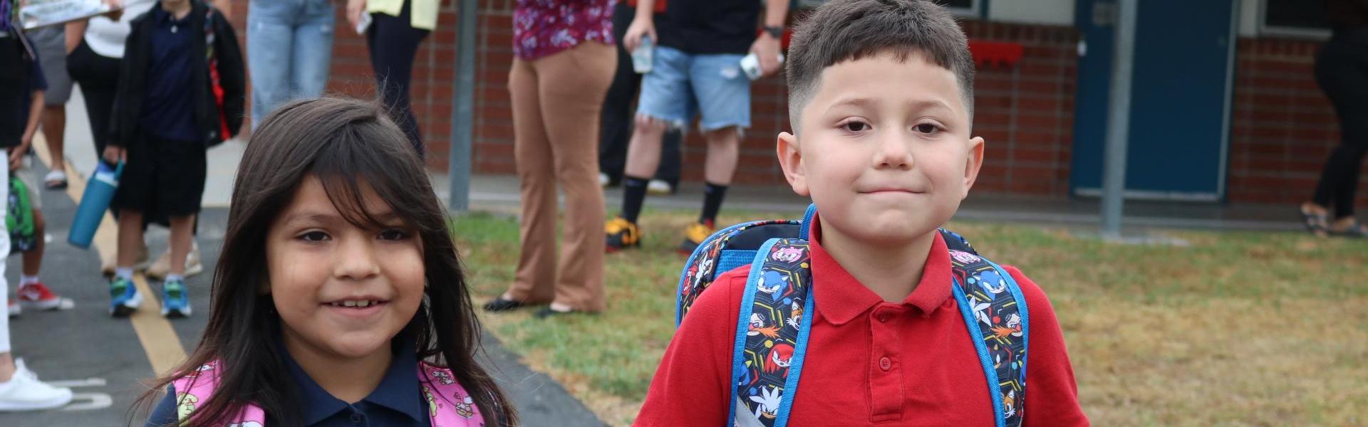 Two children walking together, smiling, wearing backpacks in a school setting.