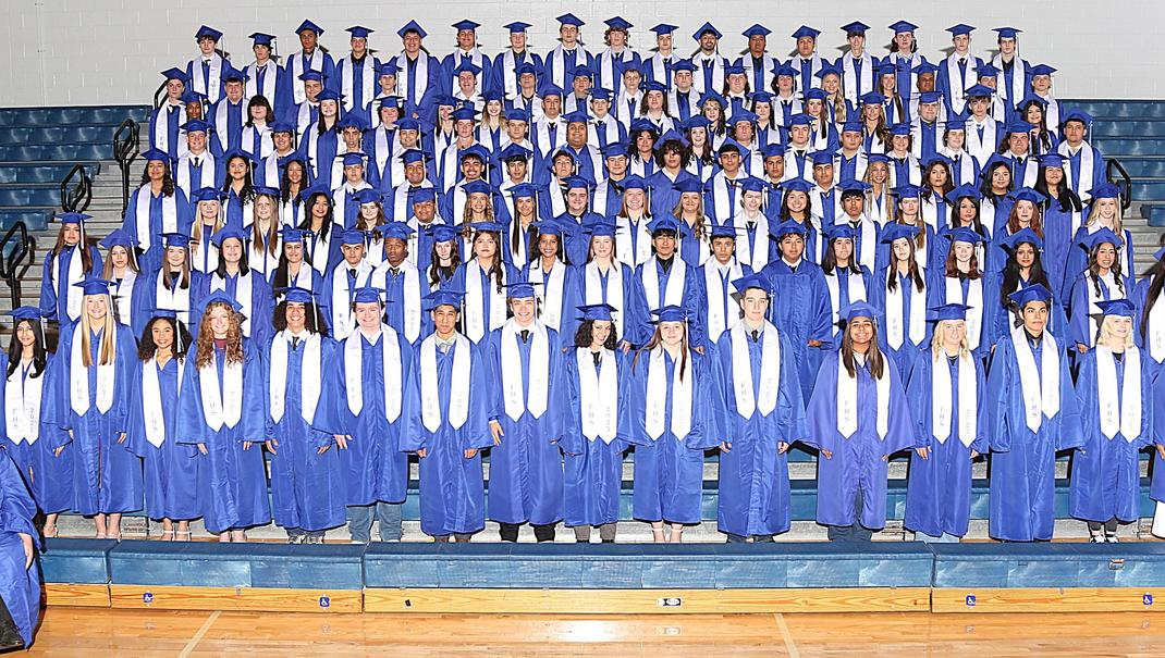 Group photo of a large graduating class in blue caps and gowns on a gymnasium stage.