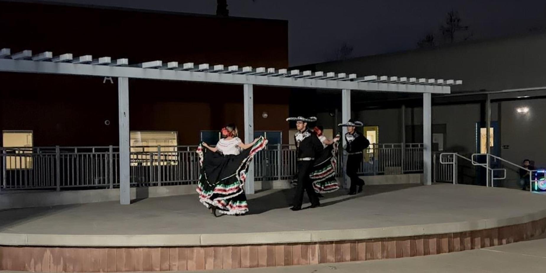 Performers in traditional attire dance on a stage at night under a pergola.