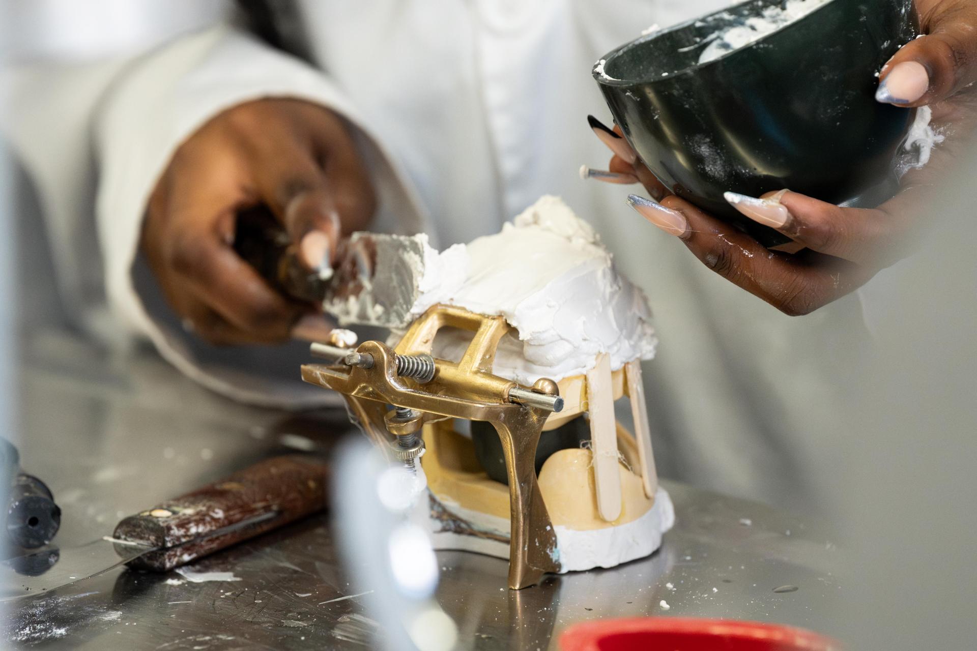 Hands apply plaster to a dental model using a bowl and tools on a worktable.