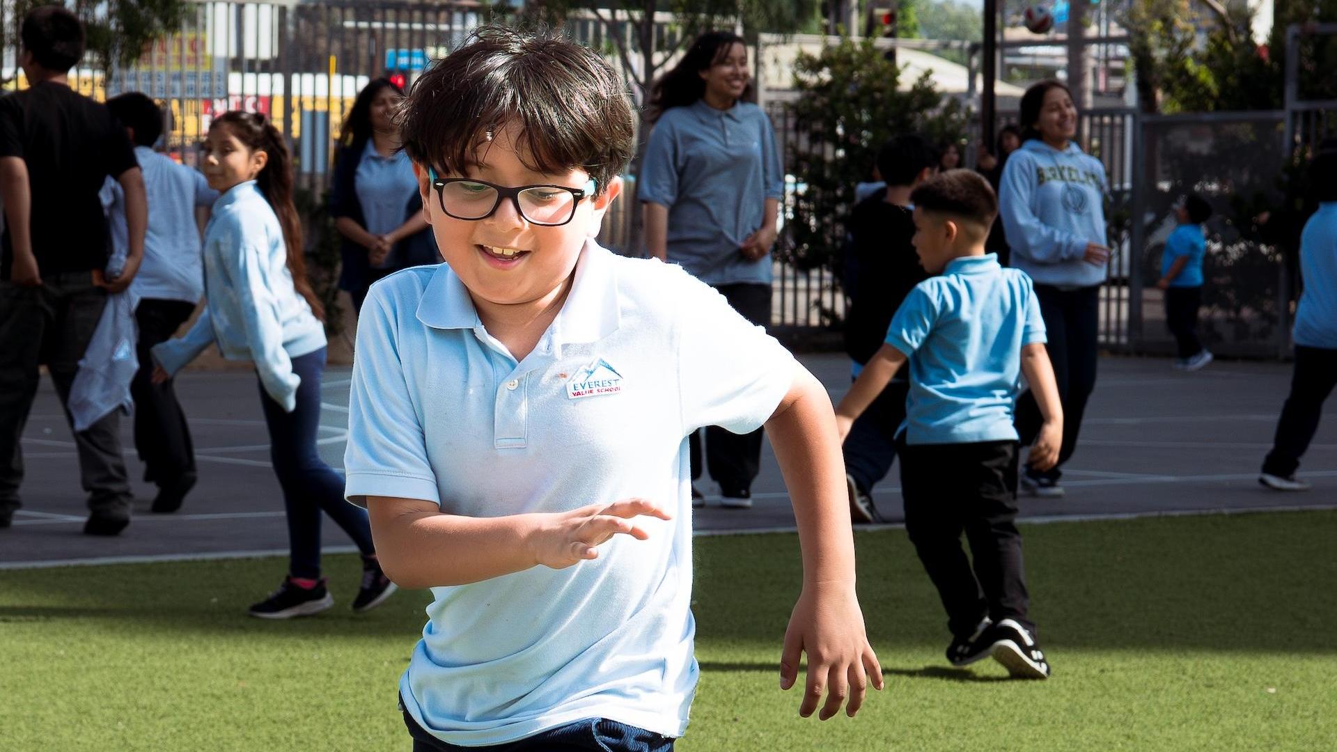 A boy running on a grassy playground wearing a light blue shirt.